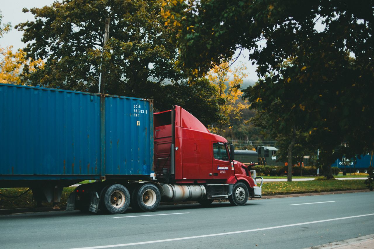 About A red truck transports a blue cargo container on a scenic, tree-lined road.